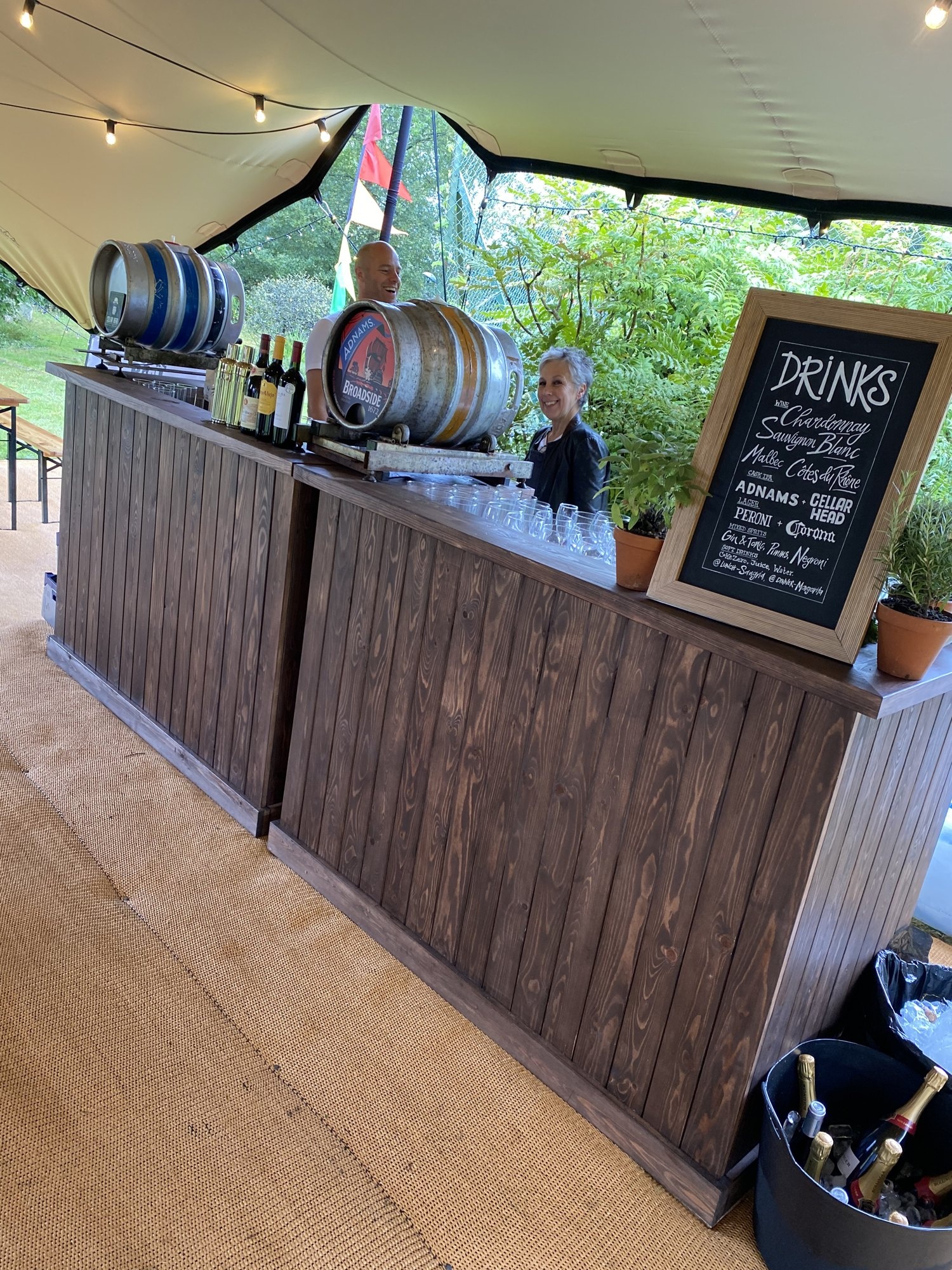 Rustic wooden event bar with drinks barrels, glassware and a chalkboard drinks menu.
