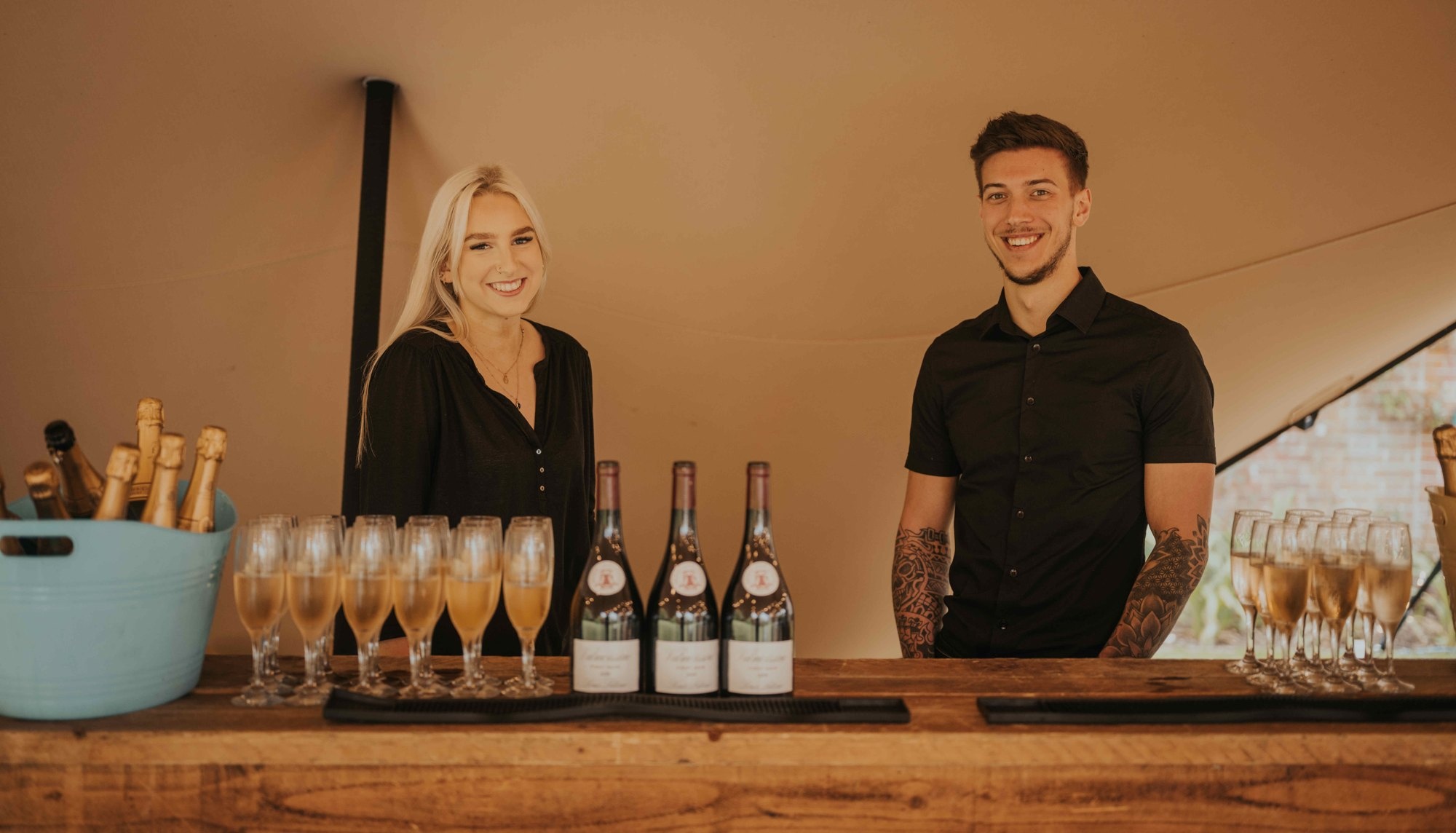 Two smiling bar staff behind a rustic wooden bar with sparkling wine and glassware.